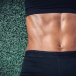 Close-up of a person's defined abdominal muscles, wearing a black sports bra and shorts, against a textured green surface.