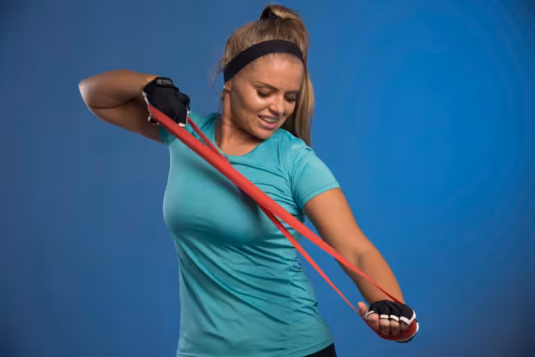 A woman wearing a black headband and teal shirt performs resistance band exercises in front of a blue wall.