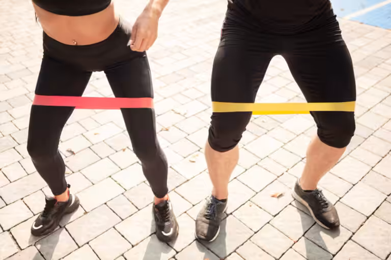 Man and woman wearing resistance bands around their thighs while squatting outdoors on a tiled surface in athletic clothing.
