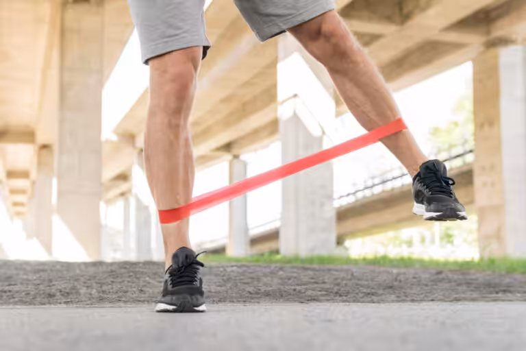 Closeup of man's legs wearing gray shorts and black sneakers performing a leg exercise with a red resistance band. under an overpass.
