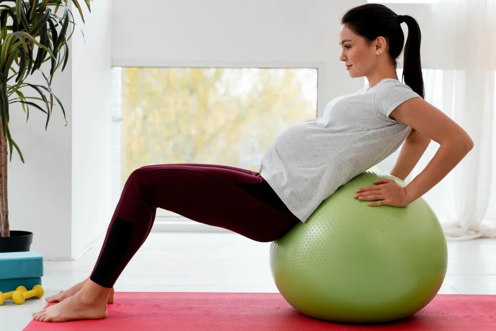 A pregnant woman in a gray shirt and dark leggings leans back on a green exercise ball on a pink mat.