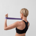 A person with short blonde hair and a black sports bra pulls a purple resistance band behind their neck.