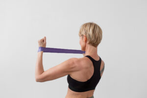 A person with short blonde hair and a black sports bra pulls a purple resistance band behind their neck.