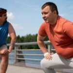Two obese men stretch outdoors. The man on the right wears an orange shirt and gray pants. The man on the left wears blue.