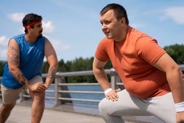 Two obese men stretch outdoors. The man on the right wears an orange shirt and gray pants. The man on the left wears blue.