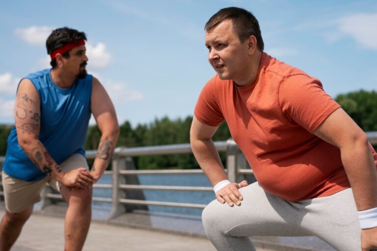 Two obese men stretch outdoors. The man on the right wears an orange shirt and gray pants. The man on the left wears blue.