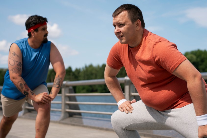 Two obese men stretch outdoors. The man on the right wears an orange shirt and gray pants. The man on the left wears blue.