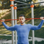 A man in a blue long-sleeve shirt holds a red resistance band behind his head at an outdoor gym.
