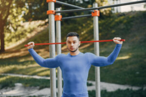 A man in a blue long-sleeve shirt holds a red resistance band behind his head at an outdoor gym.