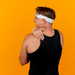 A person wearing a black tank top and white headband grasps their right shoulder in pain in front of an orange wall.