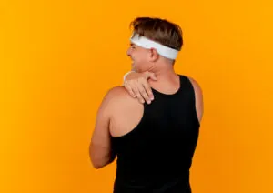 A person wearing a black tank top and white headband grasps their right shoulder in pain in front of an orange wall.