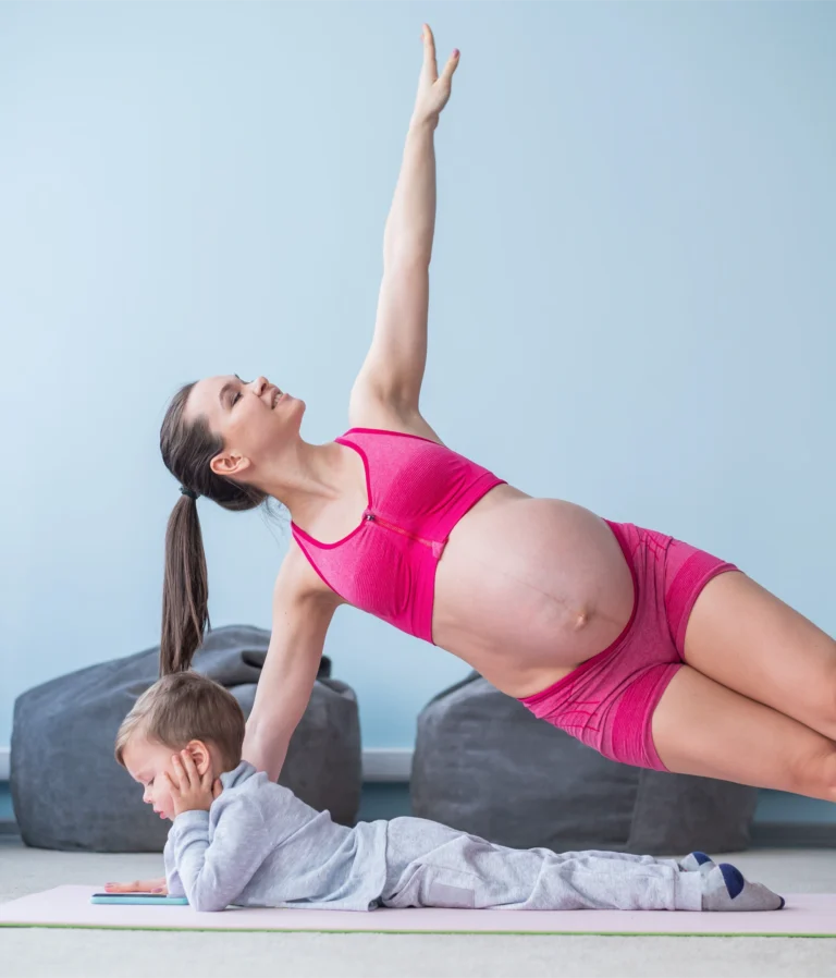 A pregnant woman in pink workout gear does a side plank. A young child lies on a mat beside her.