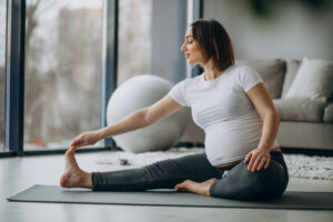A pregnant woman in a white shirt and gray pants stretches on a yoga mat indoors. Pregnancy ball, sofa, and window behind her.
