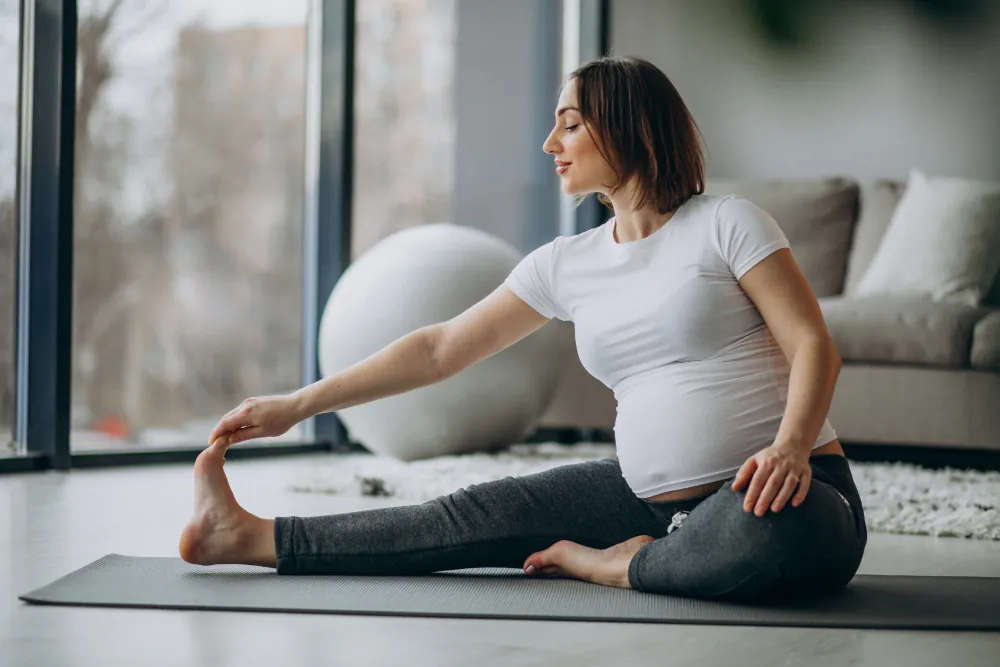 A pregnant woman in a white shirt and gray pants stretches on a yoga mat indoors. Pregnancy ball, sofa, and window behind her.