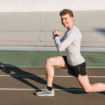 A smiling man in a gray shirt and black shorts performs a lunge on an outdoor track.