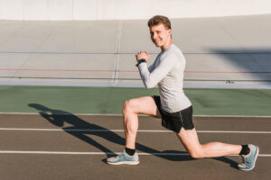 A smiling man in a gray shirt and black shorts performs a lunge on an outdoor track.