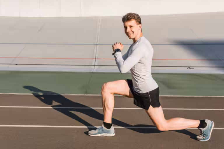 A smiling man in a gray shirt and black shorts performs a lunge on an outdoor track.