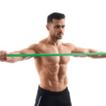 A muscular man in black shorts pulls a green resistance band horizontally across his chest.