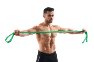 A muscular man in black shorts pulls a green resistance band horizontally across his chest.