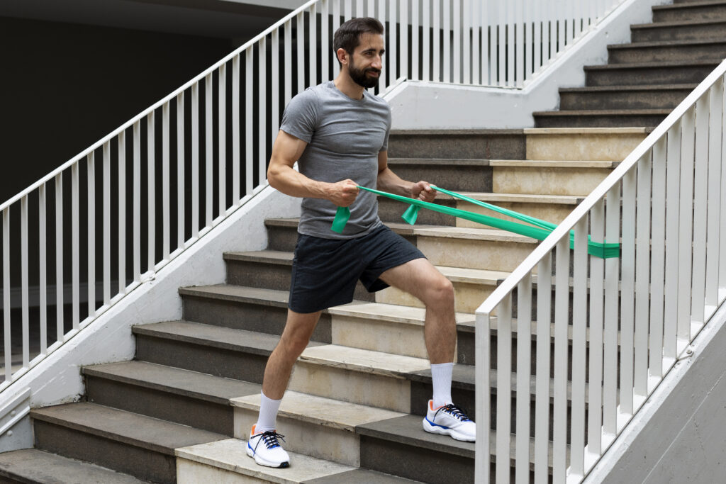 Man in a gray shirt and black shorts uses a green resistance band while exercising on outdoor stairs.