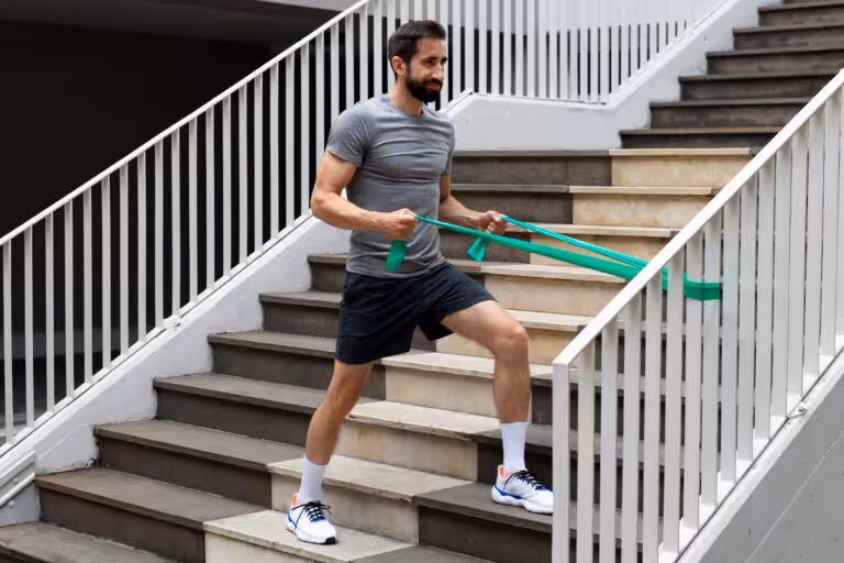 Man in a gray shirt and black shorts uses a green resistance band while exercising on outdoor stairs.