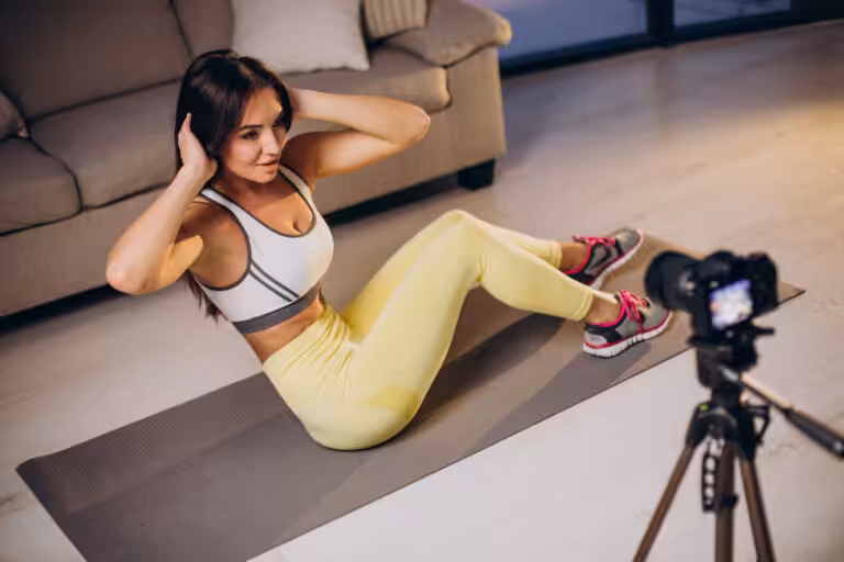 Woman in a white sports bra and yellow leggings does crunches on a gray mat, filmed by a camera on a tripod.