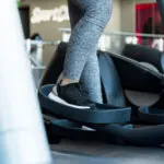A person wearing gray leggings and black sneakers stands on an elliptical machine.