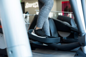 A person wearing gray leggings and black sneakers stands on an elliptical machine.