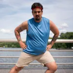 A man with a red headband and a blue tank top stretches outside next to a railing and water.