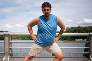 A man with a red headband and a blue tank top stretches outside next to a railing and water.