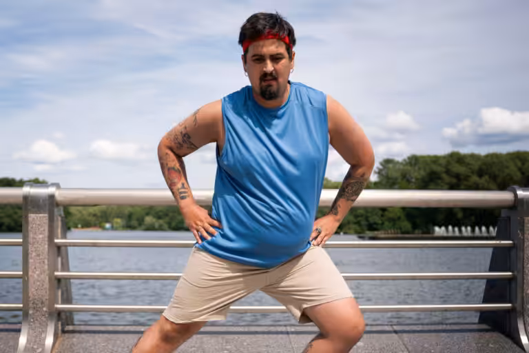 A man with a red headband and a blue tank top stretches outside next to a railing and water.