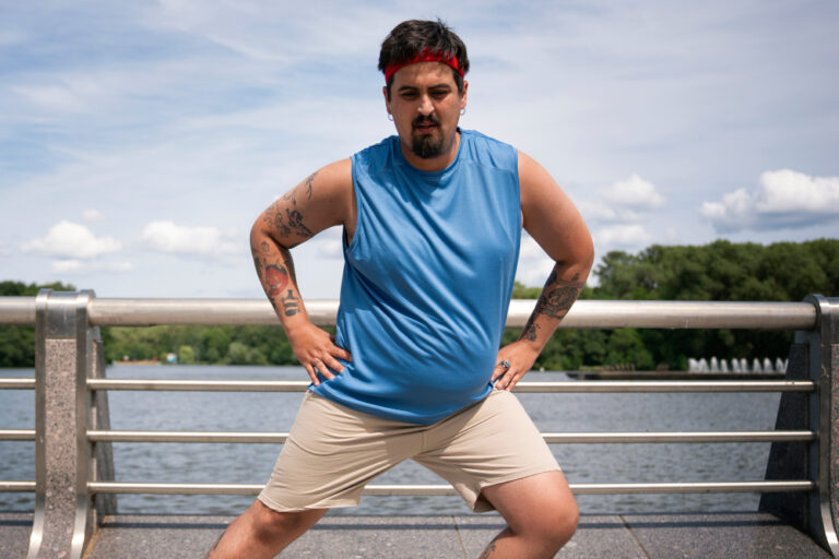 A man with a red headband and a blue tank top stretches outside next to a railing and water.