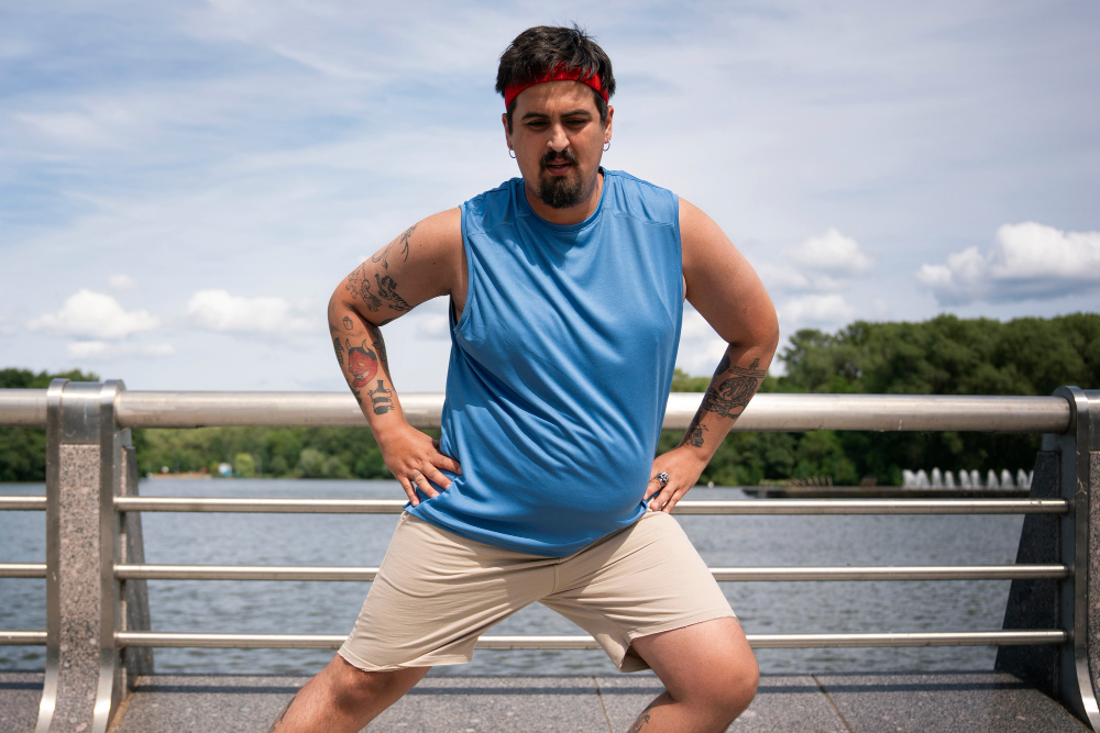 A man with a red headband and a blue tank top stretches outside next to a railing and water.