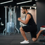 A man in black workout clothes performs a lunge with his back leg on a bench inside a gym.