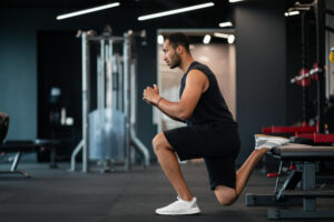 A man in black workout clothes performs a lunge with his back leg on a bench inside a gym.