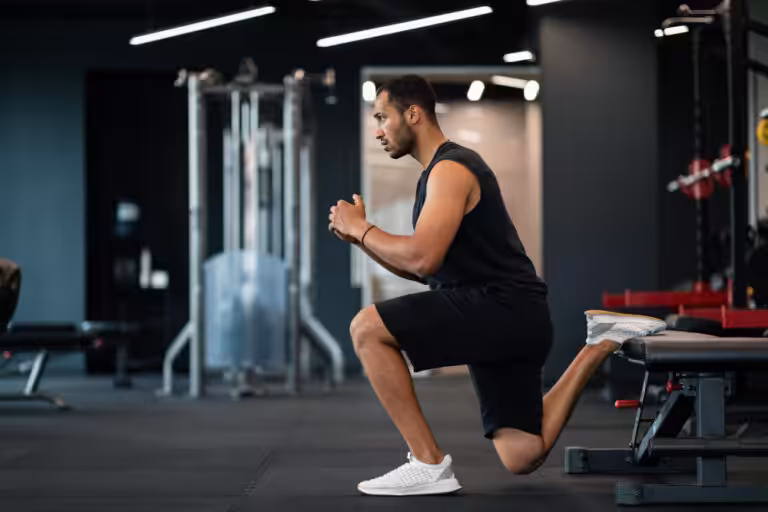 A man in black workout clothes performs a lunge with his back leg on a bench inside a gym.