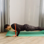 A man does pushups on a green mat, with an orange resistance band wrapped around his back.