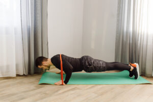 A man does pushups on a green mat, with an orange resistance band wrapped around his back.