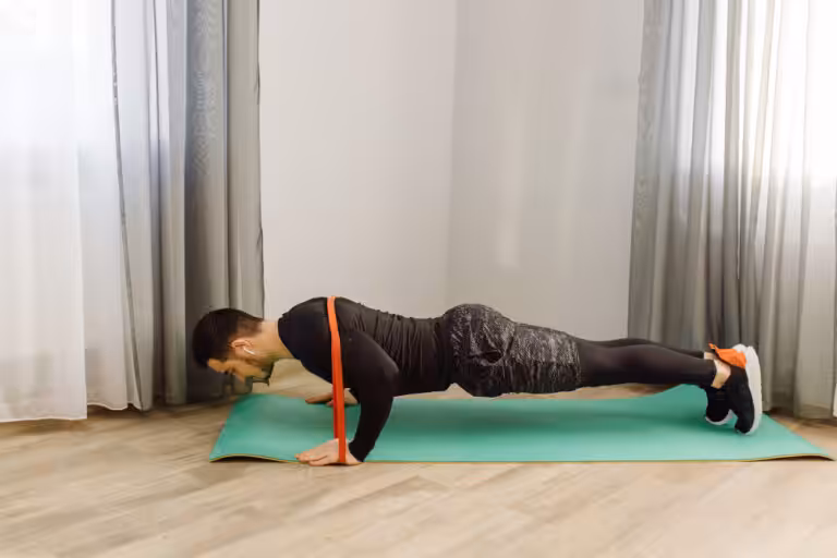 A man does pushups on a green mat, with an orange resistance band wrapped around his back.