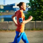 Man in red shirt and blue pants jogging outdoors with phone armband and white earbuds in golden hour lighting.