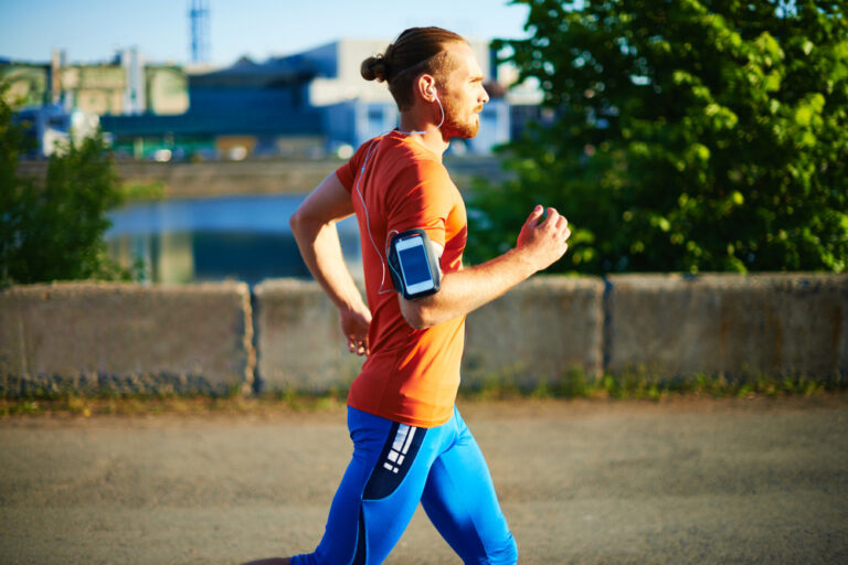 Man in red shirt and blue pants jogging outdoors with phone armband and white earbuds in golden hour lighting.