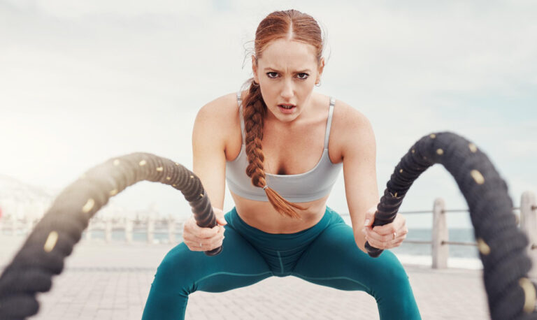Woman with braided hair in gray sports bra and teal leggings doing battle rope exercise outdoors near waterfront.