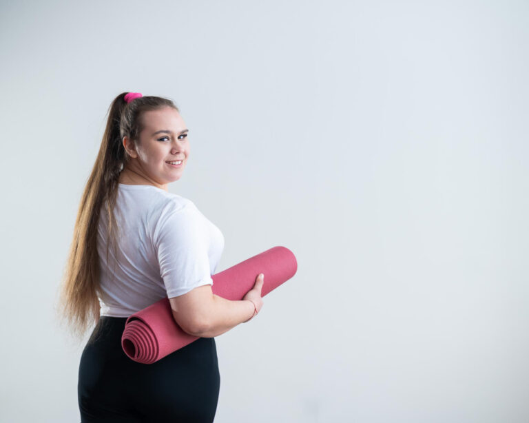 Smiling woman with long ponytail in white shirt and black pants holding rolled pink yoga mat against gray background.