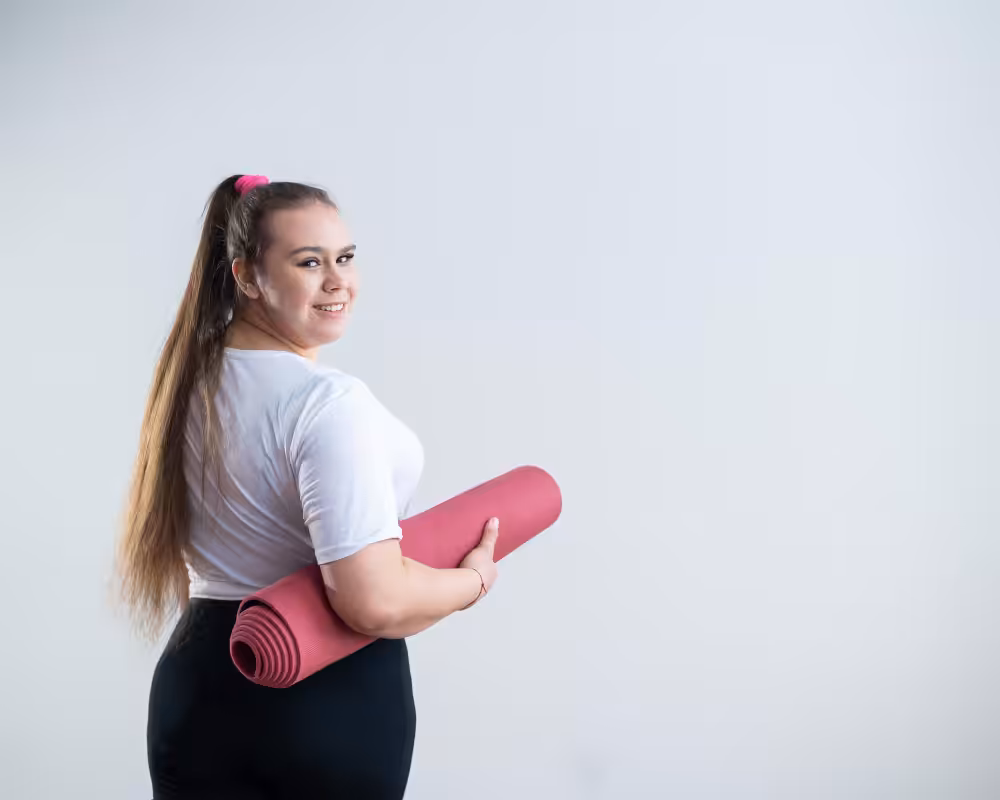 Smiling woman with long ponytail in white shirt and black pants holding rolled pink yoga mat against gray background.