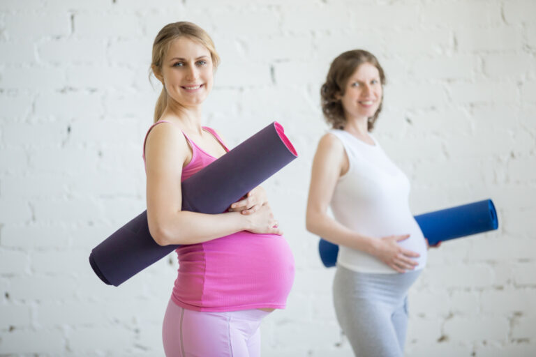 Two pregnant women smile, holding yoga mats in a bright white room.