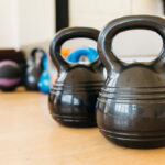Two black kettlebells with ridged handles on wooden floor with colorful fitness equipment visible in blurred background.