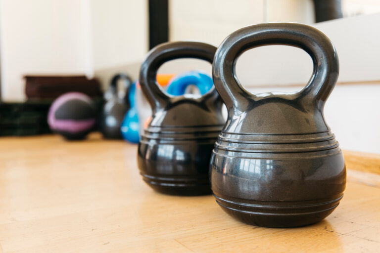 Two black kettlebells with ridged handles on wooden floor with colorful fitness equipment visible in blurred background.