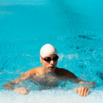 Swimmer in white cap and dark goggles performing breaststroke in bright turquoise pool water with splashing around body.