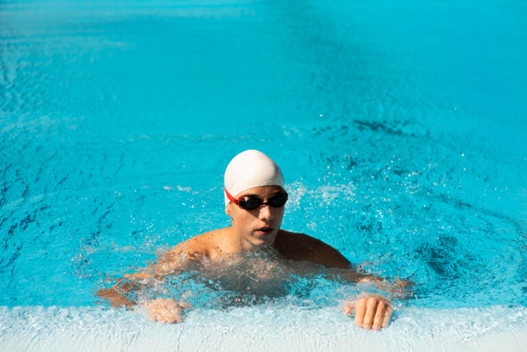 Swimmer in white cap and dark goggles performing breaststroke in bright turquoise pool water with splashing around body.