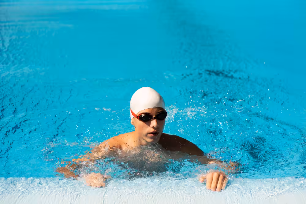 Swimmer in white cap and dark goggles performing breaststroke in bright turquoise pool water with splashing around body.